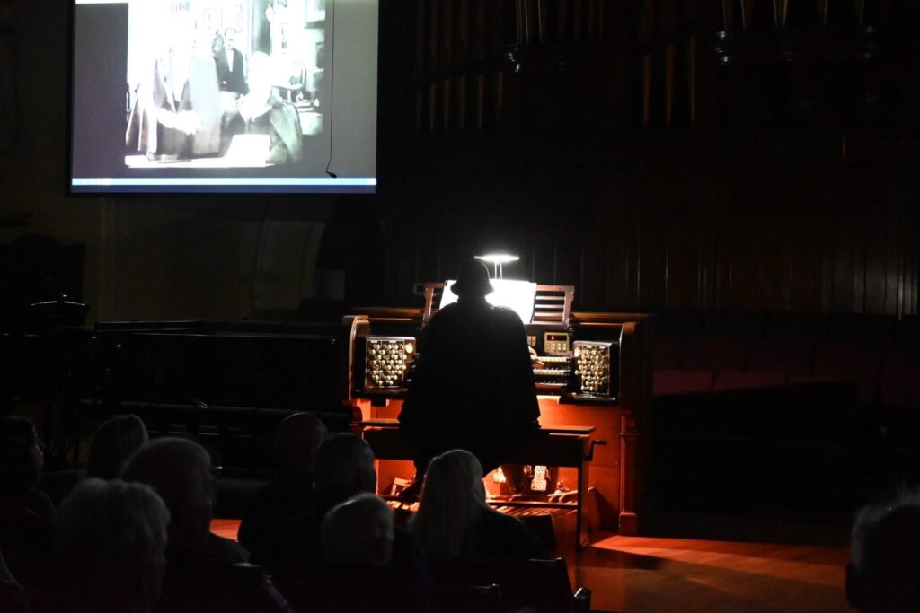 An organist sits with back to audience and black and white film running in the background.